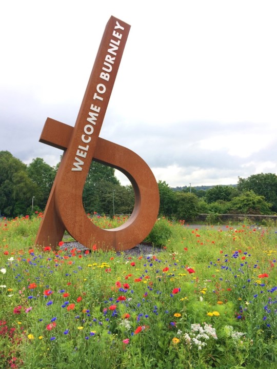 Green field with wild flowers and a metal installation that reads Welcome to Burnley