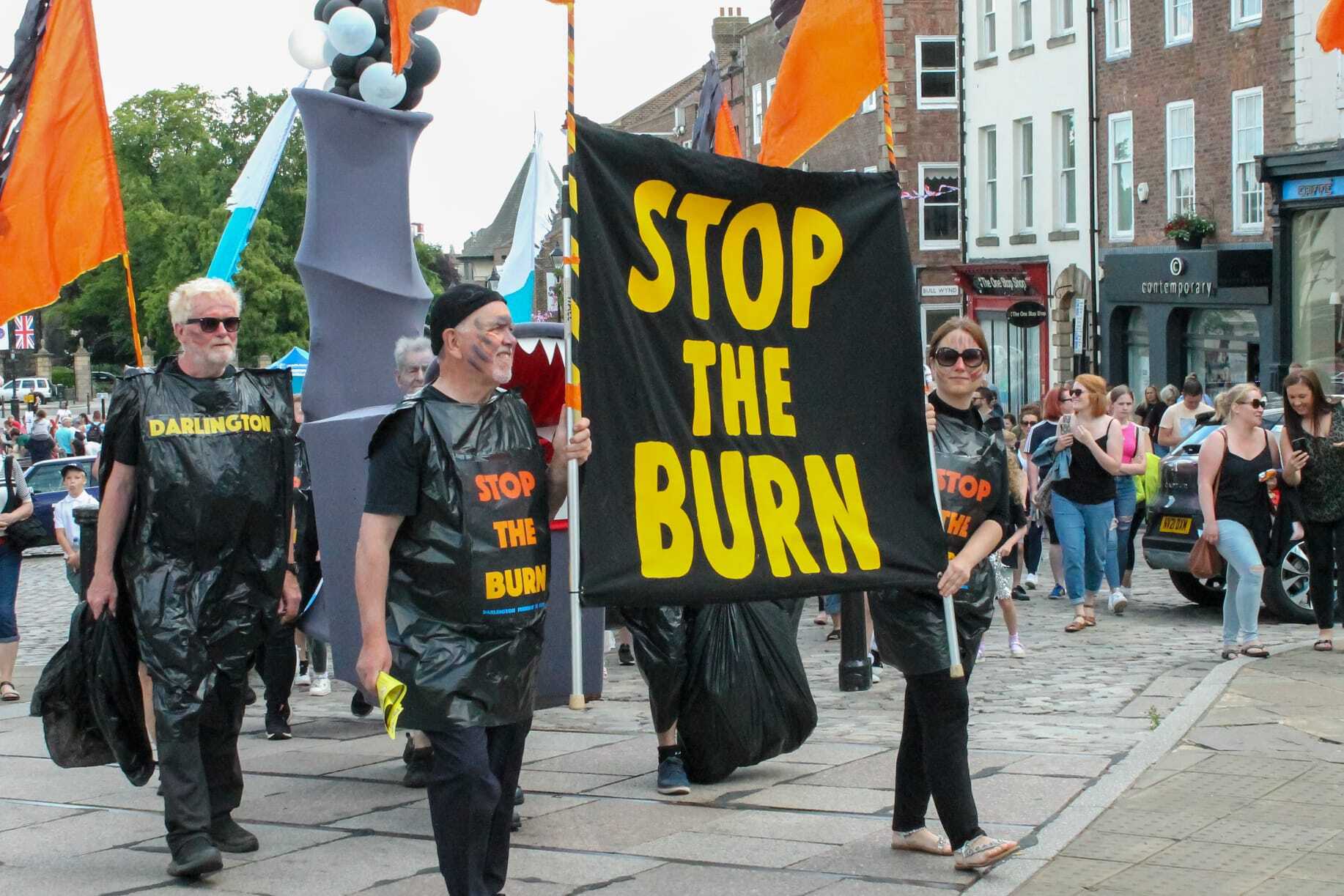 A group of people at a carnival parade carrying a model smoking incinerator and a banner saying "Stop the burn"