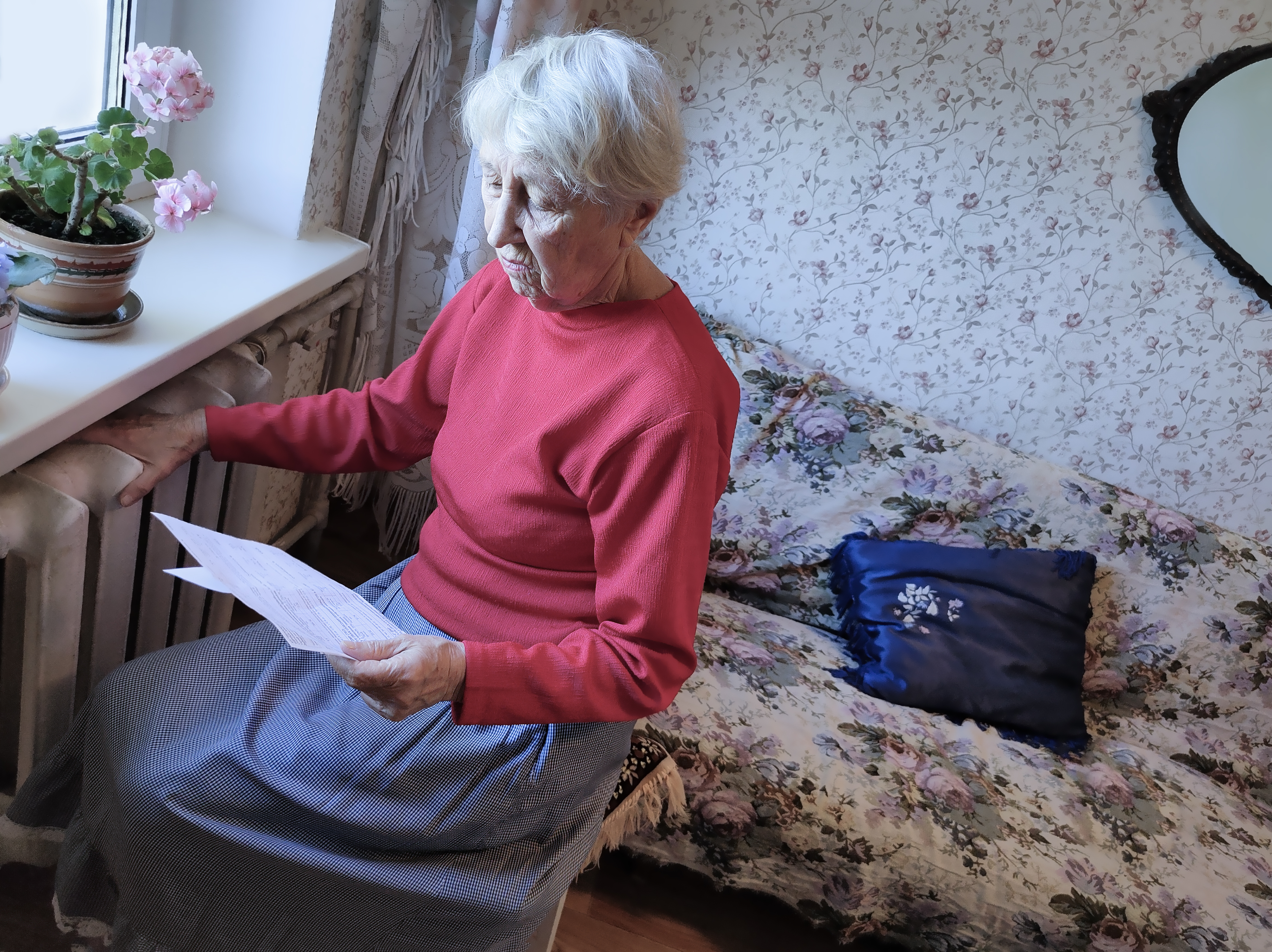 Older woman sits on her bed with her hand on a radiator as she reads a gas bill