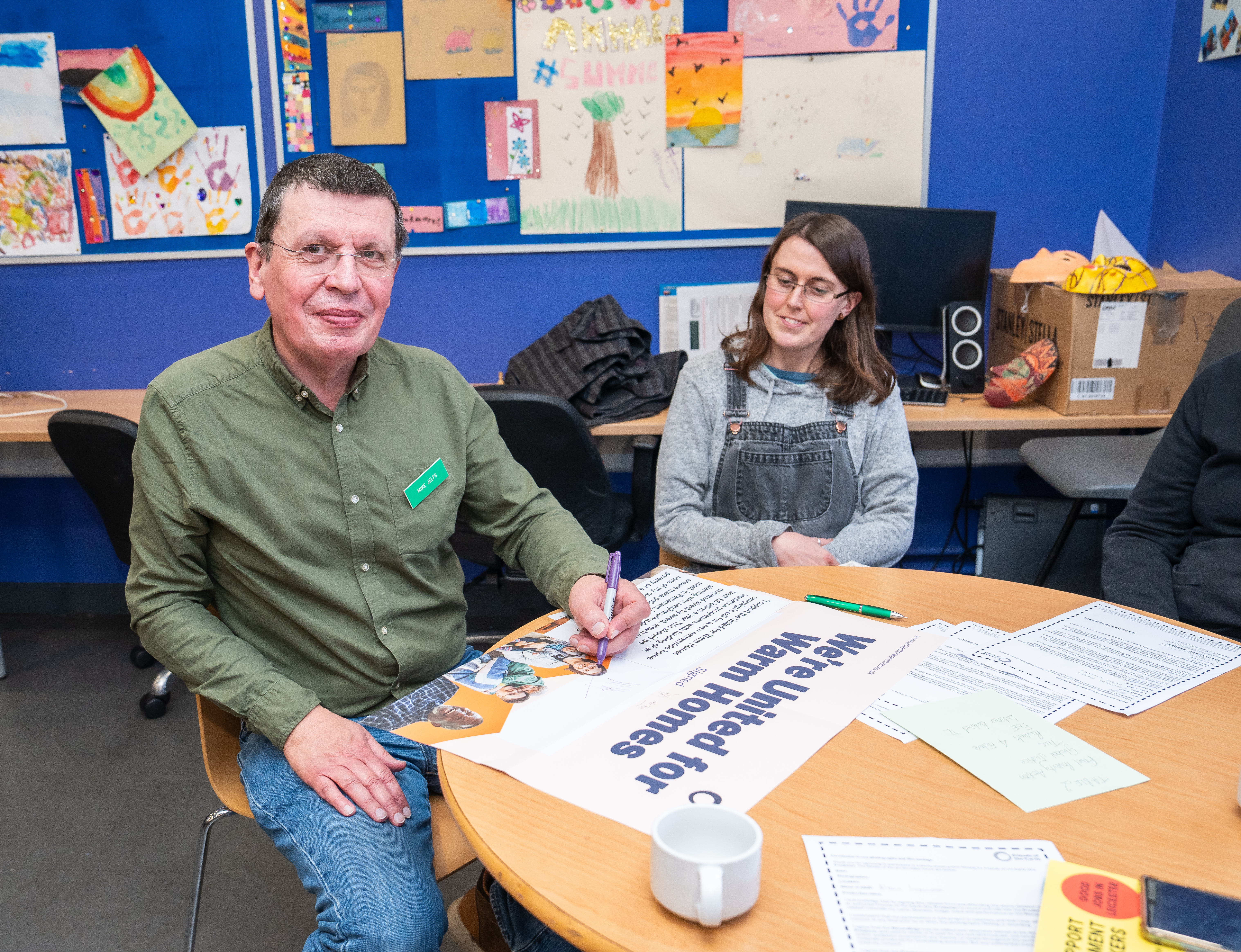 Man and woman sat at a table, the man is about to sign a partnership poster