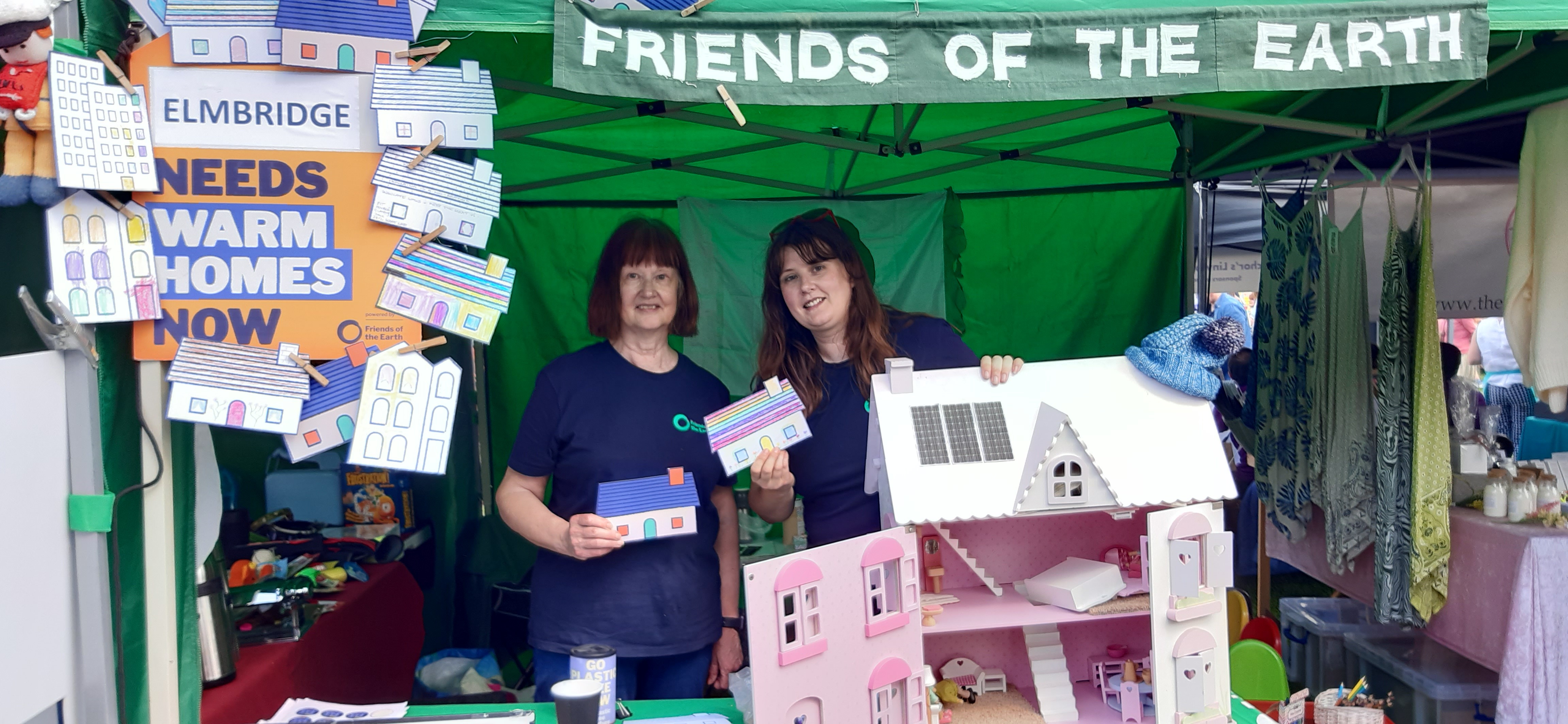 Two women stood at a stall with a dolls' house and Warm Homes postcards