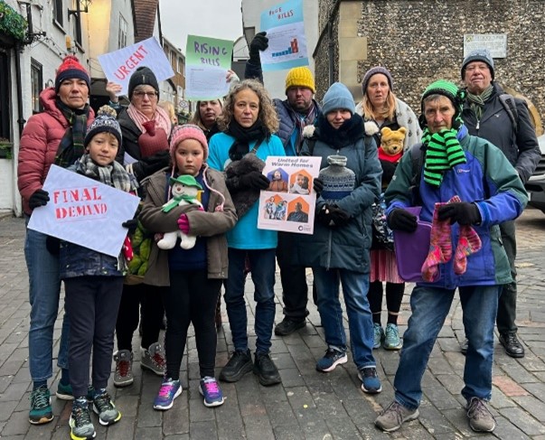 A group of people posing with scarfs, woolly hats, hot water bottles and campaign placards calling for warm homes.