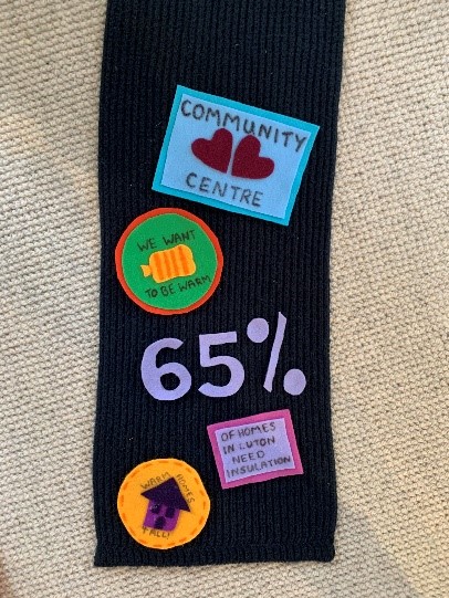 A black scarf with fabric patches attached to it, one for a community centre, one that says "We want to be warm", and one that says "65% of homes in Luton need insulation".