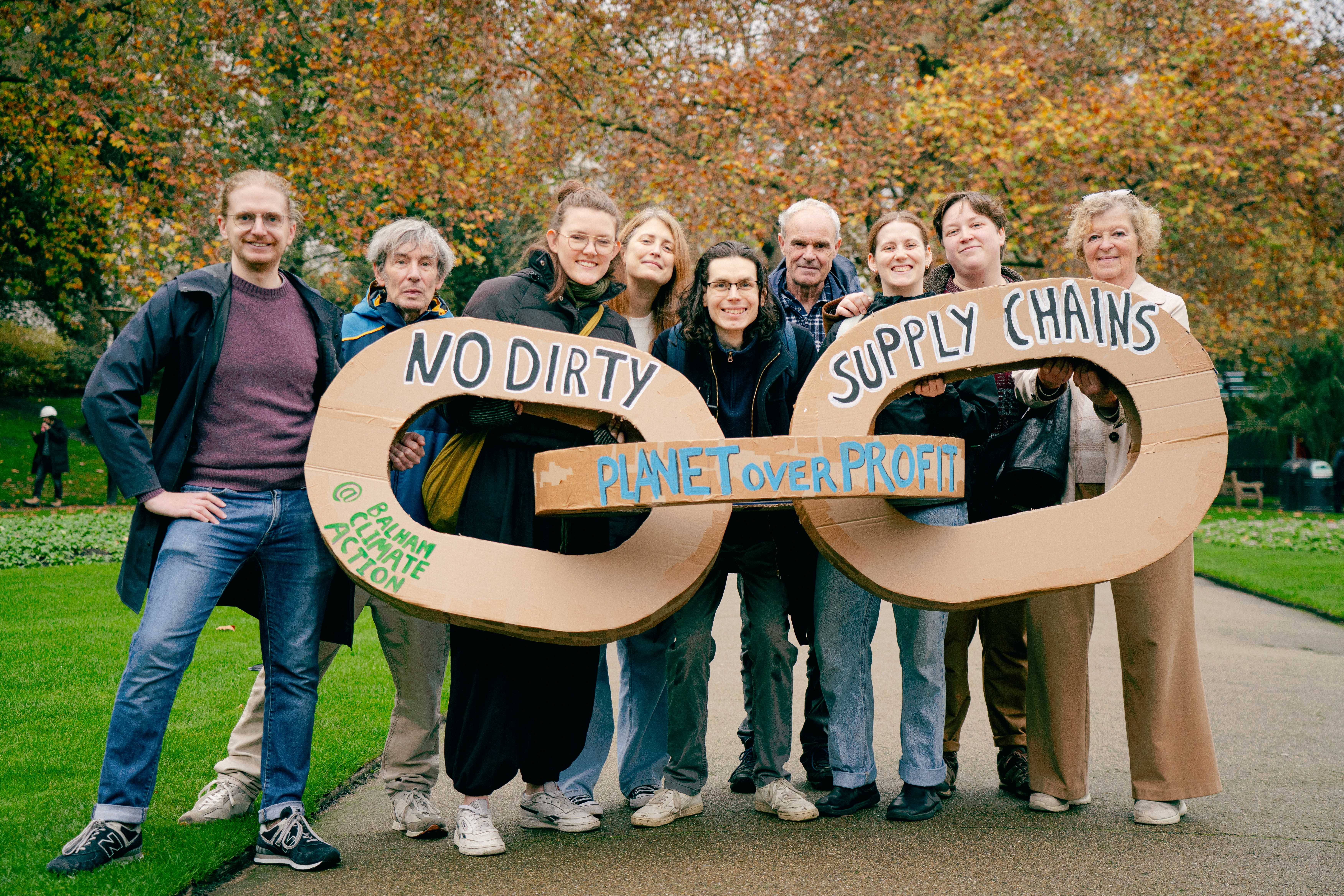 9 activists holding three giant chain links made of cardboard that say "no dirty supply chains" on a pavement in a park with autumn trees behind them 