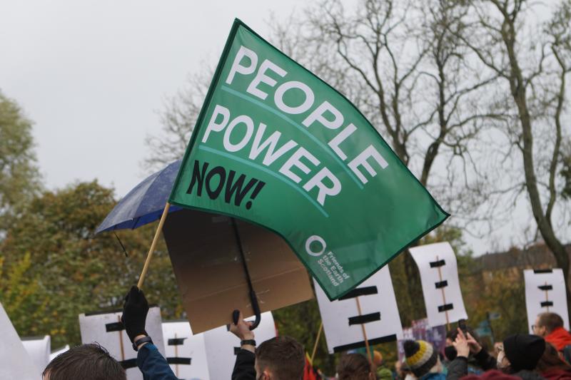 Image of protest, zoomed into hand a flag which reads 'people power now'