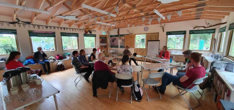 A group shot of people in an airy room, with lots of windows sitting around tables taking part in a meeting