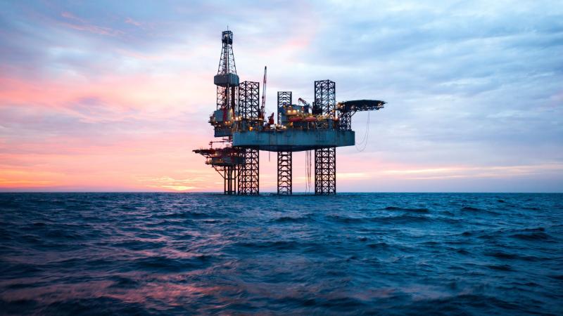 An image of an oil rig at sea. The sun is setting an the sky is a beautiful medley of colours, contrasted with the dark blue sea. It's a real shame the oil rig in the sea is so ugly.