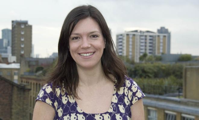 A headshot of a woman with dark hair smiling directly at the camera, among a city backdrop