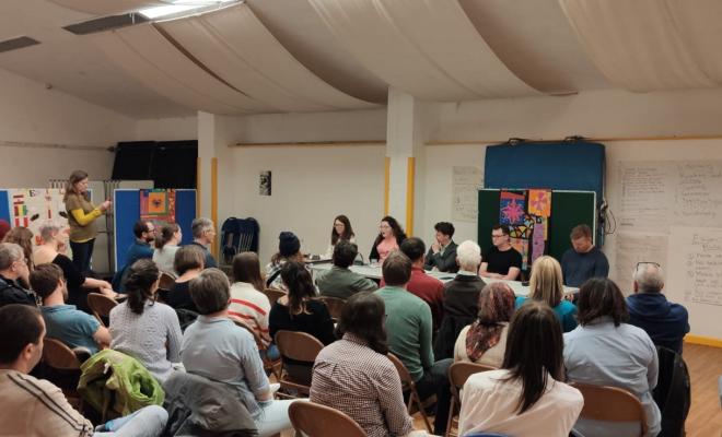 A meeting is taking place - the photo is taken so we can see the backs of the audience in chairs, listening closely to the panel of 4 people speaking