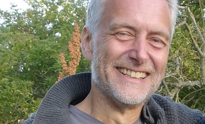 A man smiling straight at the camera, he looks warm and friendly and is in a wooded area