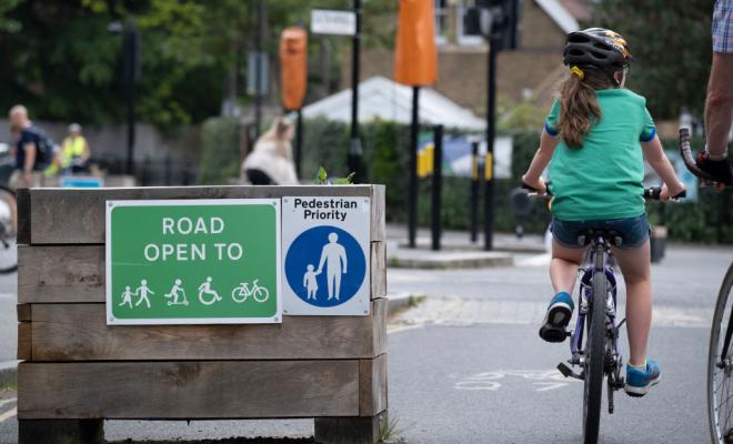 Child wearing helmet cycles past a wooden planter with signs signalling a low traffic neighbourhood