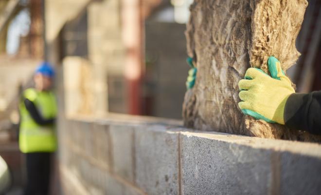 Close up of a worker's gloved hands installing cavity insulation in a wall, colleague in the background