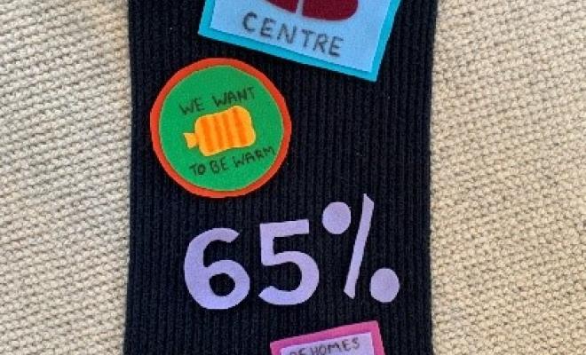 A black scarf with fabric patches attached to it, one for a community centre, one that says "We want to be warm", and one that says "65% of homes in Luton need insulation".