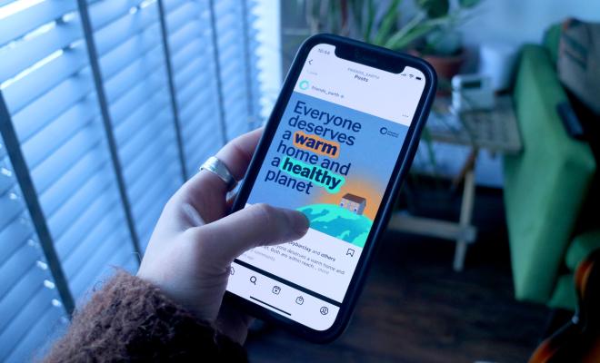 A person's hand holding their phone, showing a social media post that says "Everyone deserves a warm home and a healthy planet"