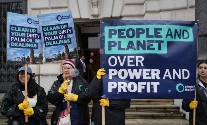 Campaigners stand outside a building holding placards and wearing yellow washing up gloves, while bubbles float in the air. The placards read: "People and planet over power and profit" and "Clean up your dirty palm oil dealings"