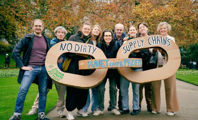 9 activists holding three giant chain links made of cardboard that say "no dirty supply chains" on a pavement in a park with autumn trees behind them 